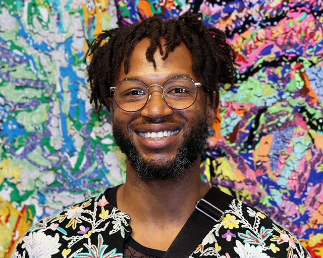 Black man with glasses, short hair, and a beard, wearing a colorful floral print jacket, smiling into the camera against a colorful and abstract backdrop