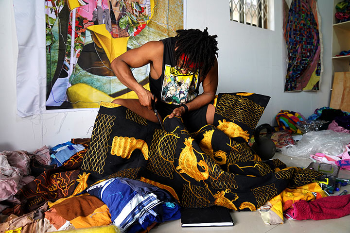 a seated Black man in a dark cutoff tee cutting ornate fabric