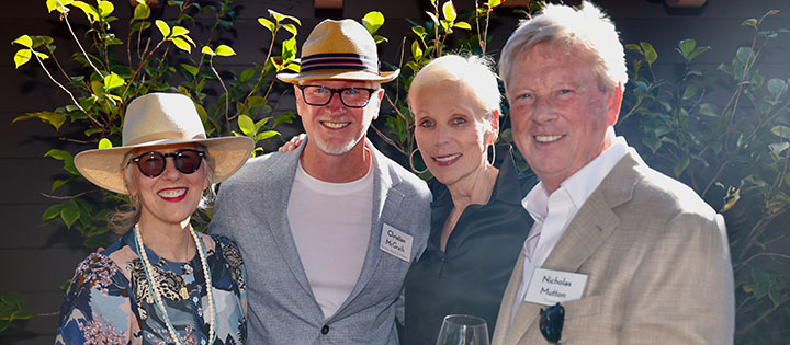 Four adults pose together outdoors, smiling at the camera. Two women and two men are dressed in stylish summer clothing and hats; one man holds a glass of white wine. Lush greenery and a building are visible in the background.