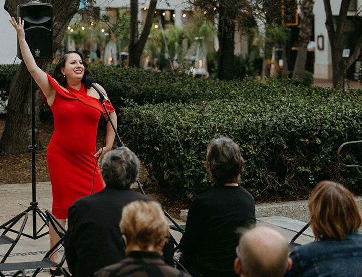 Singer in a red dress, posing with her arm raised in front of a crowd on a terrace.