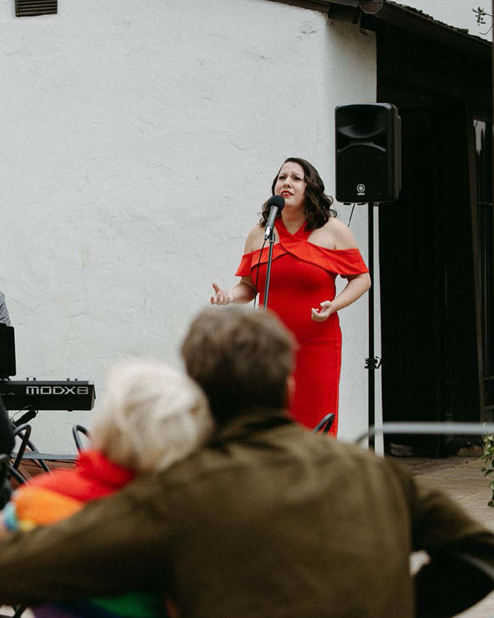Singer in a red dress, posing with her arms slightly raised in front of a crowd and microphone on a terrace.